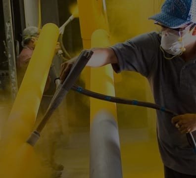 A man in blue shirt and hat sprays yellow paint on a yellow pipe for powder coating