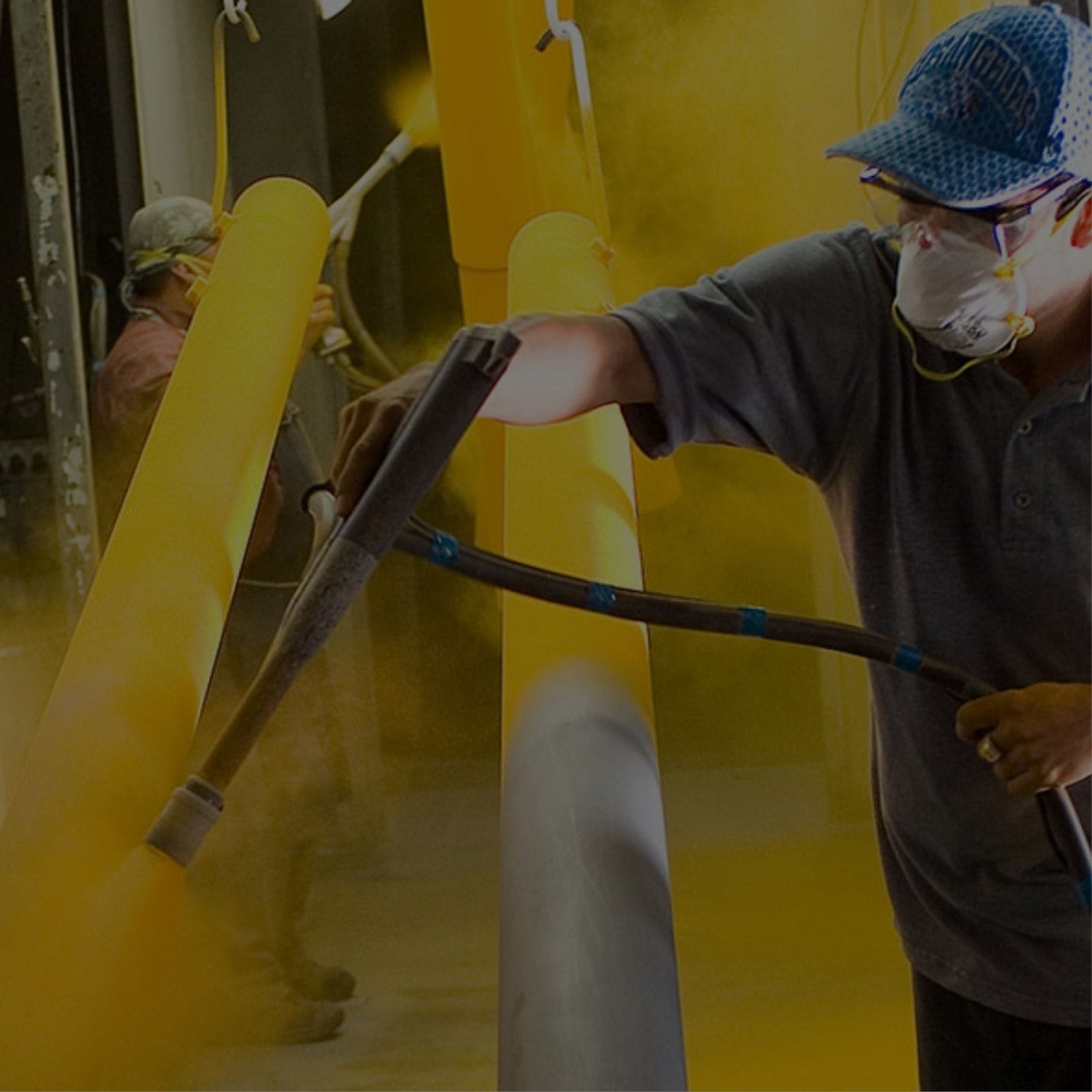 A man in blue shirt and hat sprays yellow paint on a yellow pipe for powder coating