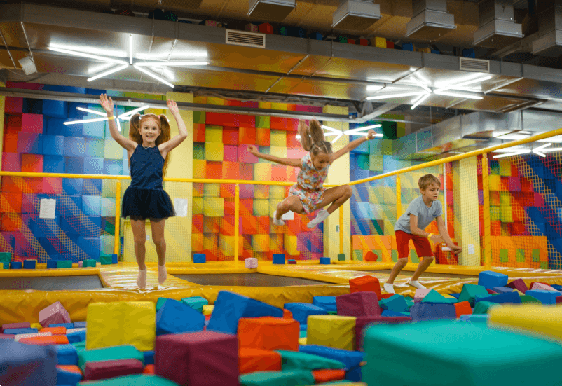 Children joyfully jumping on a trampoline in a vibrant indoor play area filled with colorful equipment.